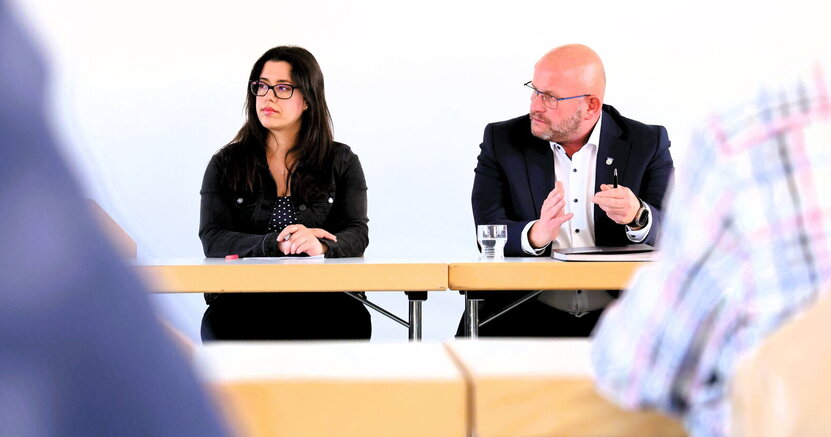 Giulia Reinhardt vom Bürgerservice und Bürgermeister Marcus Kretschmann sitzen an einem Tisch, hinter ihnen befindet sich eine weiße Wand. Der Bürgermeister gestikuliert mit den Händen.