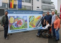 Erster Kreisbeigeordneter Adil Oyan, Erster Stadtrat Richard Kraft, Kulturbüroleiter Marco Hardy, Behindertenbeauftragte Marina Wohner sowie ihre Eltern Horst und Hannelore Wohner (v.li.) bei der Ausstellungseröffnung. Auf dem Rathausplatz stehen vor einem großen Banner mit zwei bunten Gemälden der Erste Kreisbeigeordneter Adil Oyan, Erster Stadtrat Richard Kraft, Kulturbüroleiter Marco Hardy, Behindertenbeauftragte Marina Wohner sowie ihre Eltern Horst und Hannelore Wohner (v.li.)