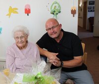 Bürgermeister Marcus Kretschmann gratulierte Elfriede Behrendt zu ihrem 103. Geburtstag. Elfriede Behrendt sitzt mit rosa Pullover an einem Tisch mit einem Geschenkkorb. Bürgermeister Marcus Kretschmann sitzt an ihrer Seite und hat den Arm auf ihre Stuhllehne gelegt.