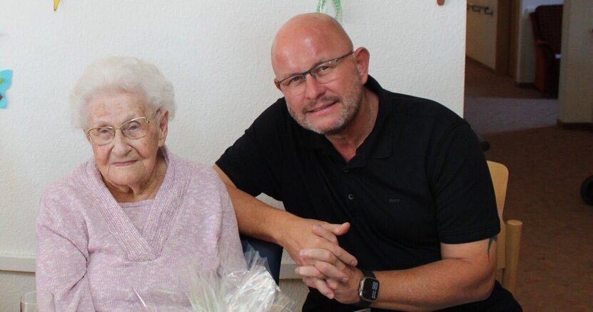 Bürgermeister Marcus Kretschmann gratulierte Elfriede Behrendt zu ihrem 103. Geburtstag. Elfriede Behrendt sitzt mit rosa Pullover an einem Tisch mit einem Geschenkkorb. Bürgermeister Marcus Kretschmann sitzt an ihrer Seite und hat den Arm auf ihre Stuhllehne gelegt.
