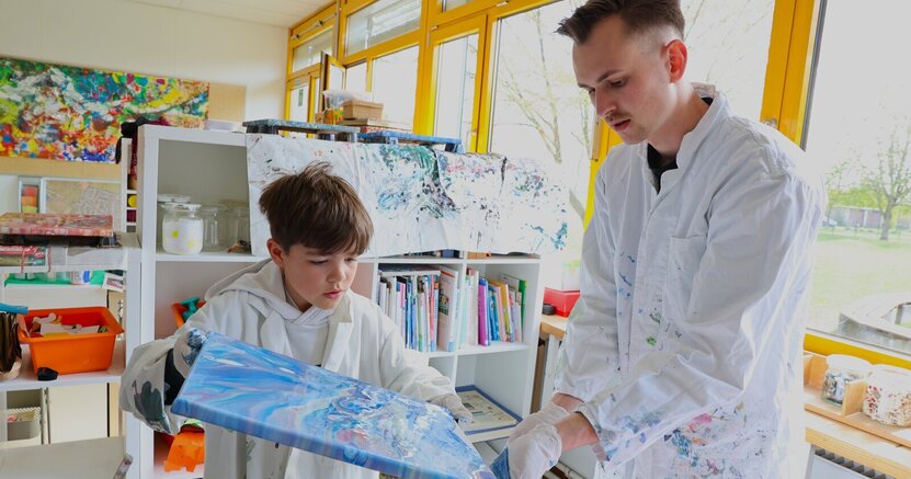 Justus malt mit der Pouring-Technik, David Kalweit von der Schuki Leeheim unterstützt ihn dabei. Eine Junge im Malerkittel kippt eine Leinwand voller Farbe, damit die Farbe verläuft. Ein Mann hilft ihm, dass nicht zu viel Farbe von dem Bild tropft.
