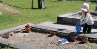 Ein Mädchen mit einer Kappe begießt eine Pflanzen in einem bemalten Topf. Er steht mit weiteren Töpfen in einem Hochbeet mit Sand.
