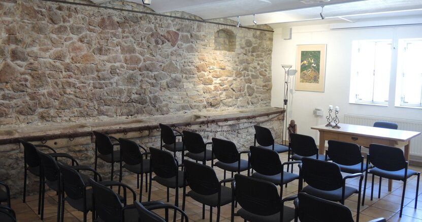 In der Kunstgalerie am Büchnerhaus stehen Stuhlreihen vor einem Tisch. Die rückwärtige Längswand besteht aus großen alten Steinen.