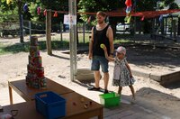 Ein kleines Mädchen im Sommerkleid und mit Sonnenhut wirft gerade einen Ball auf eine Dosen-Pyramide, die auf einem Tisch vor ihr steht. Neben ihr steht ein Mann und schaut zu.