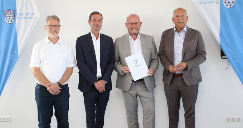 Die Bürgermeister Thomas Raschel (Stockstadt), Marcus Rahner (Biebesheim) und Marcus Kretschmann (Riedstadt) mit dem Hessischen Innenminister Roman Poseck (v.re.) bei der Bescheidübergabe im Riedstädter Rathaus. Im Sitzungssaal des Riedstädter Rathauses stehen der Bürgermeister von Stockstadt Thomas Raschel, der Bürgermeister von Biebesheim Marcus Rahner, der Bürgermeister von Riedstadt Marcus Kretschmann und der Hessische Innenminister Roman Poseck nebeneinander (von links nach rechts). Bürgermeister Kretschmann hält eine Mappe mit der Aufschrift "Hessen" in der Hand. Links und rechts von den Herren sind Ständer mit dem blau-weißen Wappen von Riedstadt sowie dem Schriftzug '"Riedstadt. Büchnerstadt" in Blau zu sehen.