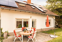 Zu sehen ist ein Paar, dass auf einer Terrasse vor ihrem Haus sitzt. Auf dem Dach ist eine PV-Anlage.
