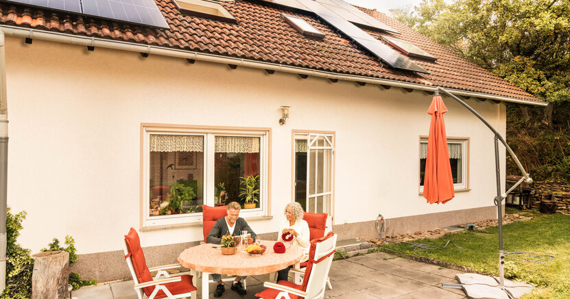 Zu sehen ist ein Paar, dass auf einer Terrasse vor ihrem Haus sitzt. Auf dem Dach ist eine PV-Anlage.
