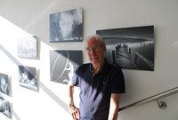 Bernd Susenburger steht vor einigen seiner Schwarz-Weiß-Fotografien, die an Wänden des Treppenhauses im Rathaus hängen. Das Bild rechts von ihm zeigt einen Steg, der ins Wasser eines Sees führt, links ist unter anderem ein Bild von einer knienden Frau zu sehen, die mit ihren Armen hinter sich einen großen Reif hält.
