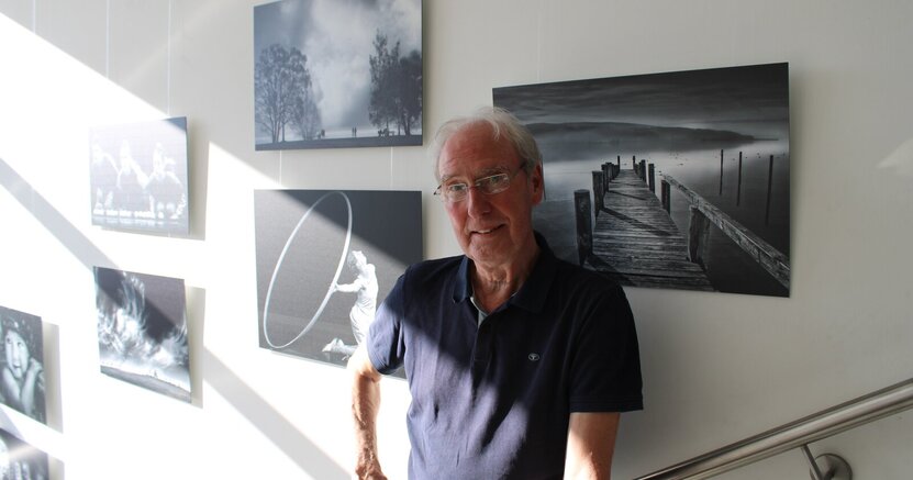 Bernd Susenburger steht vor einigen seiner Schwarz-Weiß-Fotografien, die an Wänden des Treppenhauses im Rathaus hängen. Das Bild rechts von ihm zeigt einen Steg, der ins Wasser eines Sees führt, links ist unter anderem ein Bild von einer knienden Frau zu sehen, die mit ihren Armen hinter sich einen großen Reif hält.