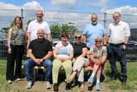 Auf der neuen Bank sitzenBürgermeister Marcus Kretschmann, Katja Glock, Ilona Jahnke und Ursula Fraikin (v.li.), daneben und dahinter stehen Sandra Seum, Mathias Lachmann, Gaston Wolfsturm und Michael Fraikin (v.li.). Im Hintergrund sind ein Zaun und dahinter Autos zu sehen.