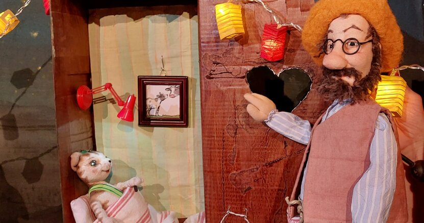 Eine Handpuppe, die den Kater Findus darstellt, liegt mit Bettdecke und Kissen in einem Klohäuschen. Die Petersson-Puppe steht davor.