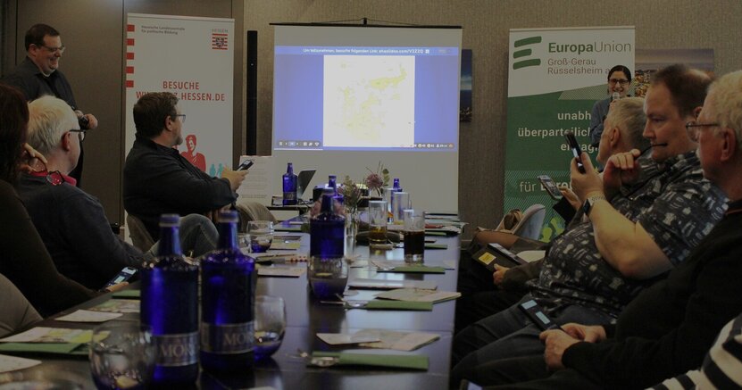 An einem langen Tisch sitzen Menschen mit ihren Smartphones. Auf einer Leinwand ist eine Frage des Pub Quiz zu sehen. Links und rechts davon stehen die Moderatoren Andreas Christ und Dr. Birgit Bujard mit Mikrofonen in der Hand.