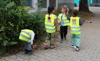Mehrere Kindergartenkinder haben gelbe Warnwesten an und stehen am Rande eines Fußwegs. Ein Junge hält eine Chipsdose hoch. Ein anderes Kind bückt sich gerade und ein weiteres hält eine Greifzange in der Hand.