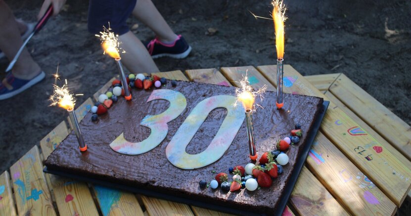 Einen Geburtstagskuchen gab es beim großen Spiel- und Spaßfest zum 30jährigen Jubiläum der Kita Thomas-Mann-Platz. Auf einen Gartentisch steht eine viereckige Schokotorte mit der Aufschrift "30" und brennenden Wunderkerzen