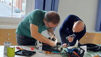 Gemeinschaftlich gewerkelt wird beim Reparatur-Café im alten Rathaus Crumstadt. Zwei Männer beugen sich über ein Gerät, das auf einem Tisch liegt. Der eine hält eine Taschenlampe, der andere schraubt etwas.