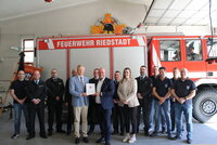 In der Fahrzeughalle der Freiwilligen Feuerwehr Erfelden stehen in einer langen Reihe vor einem Feuerwehrwagen mit der Aufschrift "Feuerwehr Riedstadt" Feuerwehrleute in Uniform. Vor ihnen überreicht Innenstaatsssekretär Martin Rößler den Zuwendungsbescheid an Bürgermeister Marcus Kretschmann.