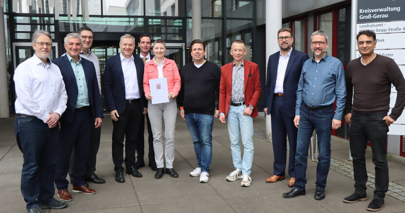 Vor dem Landratsamt stehen v.l.n.r. die Bürgermeister Thomas Raschel (Stockstadt am Rhein), Marcus Merkel (Büttelborn), Jörg Rüddenklau (Groß-Gerau), Peter Burger (Gernsheim) und Marcus Rahner (Biebesheim am Rhein), Marion Götz (Leiterin IKZ-Lenkungsgruppe), Bürgermeister Marc Friedrich (Nauheim), Landrat Thomas Will, die Bürgermeister Karsten Groß (Mörfelden-Walldorf) und Thorsten Siehr (Ginsheim-Gustavsburg) sowie Erster Kreisbeigeordneter Adil Oyan. Marion Götz hält den Förderbescheid in der Hand.