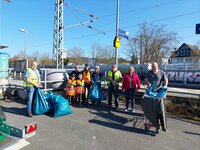 Kinder und zum Teil auch Erwachsene haben Warnwesten an und halten Greifzangen in der Hand. Einige Erwachsene halten Müllsäcke in der Hand. Sie stehen vor dem Eingang zum Bahnhof Wolfskehlen, im Hintergrund ist ein Bahnsteig zu sehen.