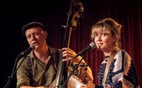 Das Duo "Stereo Naked" steht auf einer Bühne. Pierce Black hält einen Kontrabass und singt in ein vor ihme stehendes Mikrofon. Julia Zech steht neben ihm mit einer Gitarre und singt ebenfalls in ein Mikrofon.