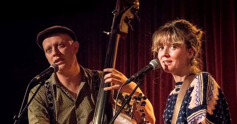 Stereo Naked (Pierce Black und Julia Zech) sorgt beim Frühschoppen am Sonntag unter den Linden für musikalische Stimmung. Das Duo "Stereo Naked" steht auf einer Bühne. Pierce Black hält einen Kontrabass und singt in ein vor ihme stehendes Mikrofon. Julia Zech steht neben ihm mit einer Gitarre und singt ebenfalls in ein Mikrofon.