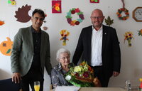 Erster Kreisbeigeordneter Adil Oyan (links) und Bürgermeister Marcus Kretschmann stehen neben Elfriede Behrendt. Sie sitzt mit einem Blumenstrauß auf einem Stuhl.