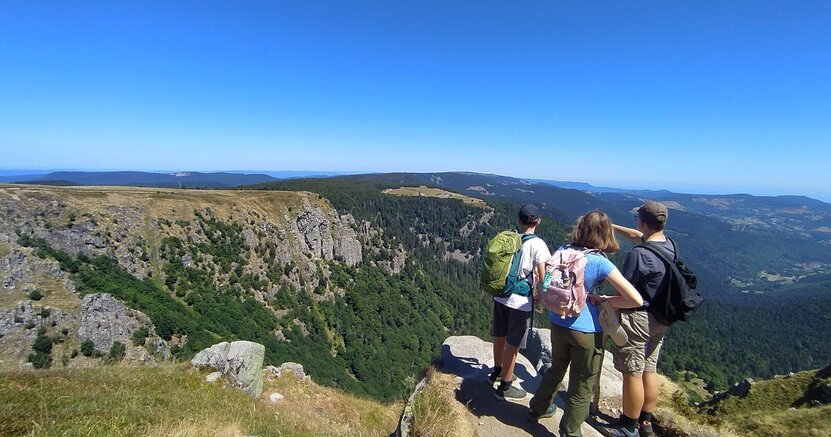 Jugendliche genießen den Ausblick bei einer Wandertour. Drei Jugendliche stehen auf einem Berg und schauen auf die Berglandschaft und den blauen Himmel.