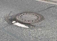 Schachtabdeckungen müssen saniert werden. Abgebildet ist eine runde Schachtabdeckung mit einer Umrandung mit Löchern aus Metall. Sie befindet sich inmitten einer Straße mit Asphaltdecke.