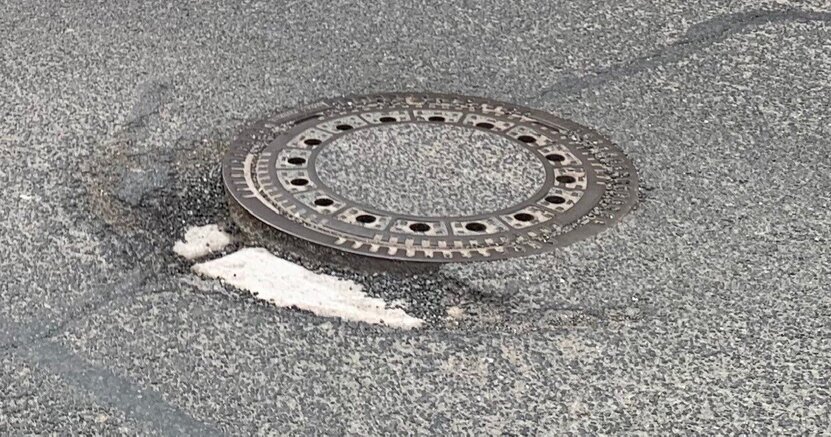 Abgebildet ist eine runde Schachtabdeckung mit einer Umrandung mit Löchern aus Metall. Sie befindet sich inmitten einer Straße mit Asphaltdecke.