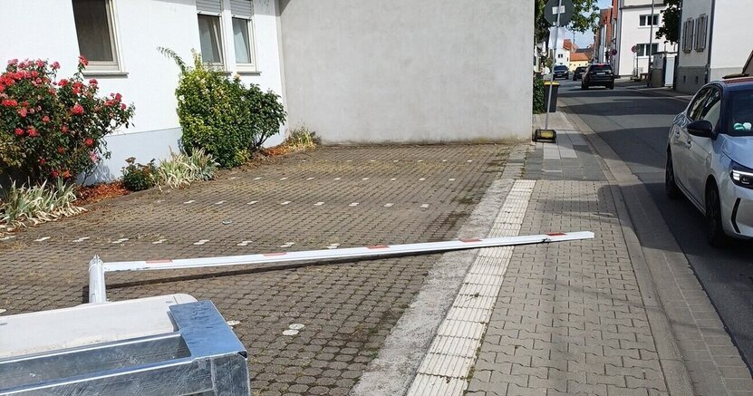 Auf dem Gehweg liegt im Vordergrund der stählerne Standfuß des Höhenbegrenzers. Dahinter liegt der rot-weiß gestreifte Höhenbegrenzer. Er sieht aus wie ein halber Rahmen.