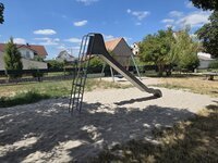 Auf dem Spielplatz Schulstraße wurde gemäß Spielplatzkonzept eine neue Rutsche aufgebaut. Auf dem Spielplatz Schulstraße steht eine neue Rutsche auf einem Sandfeld. Im Hintergrund ist noch eine Rasenfläche und dahinter Häuser zu sehen.