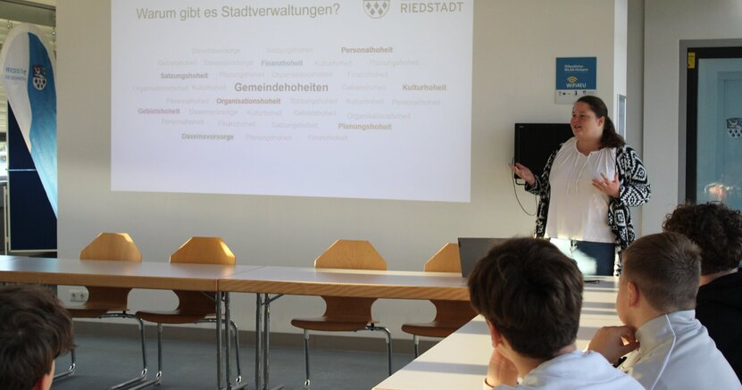 Julia Galley steht vor einer Wand im Sitzungssaal. Neben ihr wird eine Präsentation zu den Aufgaben der Stadtverwaltung an der Wand abgebildet.