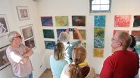 An der Wand hängen bunte Gemälde. Eine Frau steht davor und fotografiert ein Werk mit ihrem Handy. Andere Erwachsene stehen drum herum und schauen zu.