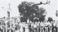 Auf der schwarz-weiß-Fotografie stehen Menschen in Badesachen um ein Schwimmbecken. Vom Sprungbrett macht ein junger Mann einen Kopfsprung. Dahinter steht ein Mann im Anzug.