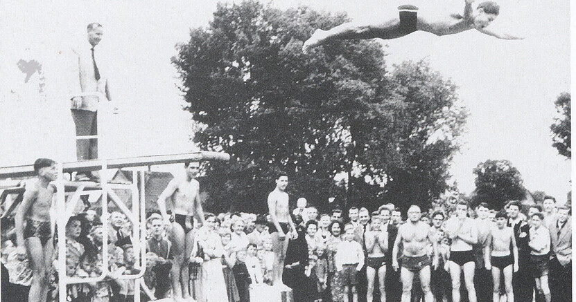 Auf der schwarz-weiß-Fotografie stehen Menschen in Badesachen um ein Schwimmbecken. Vom Sprungbrett macht ein junger Mann einen Kopfsprung. Dahinter steht ein Mann im Anzug.