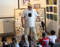 Mit der Keule wurde bei den Höhlenmenschen geregelt, wer Bestimmer ist, zeigt Forscher Dr. Gitterbaum. Dr. Gitterbaum steht mit Tropenhelm und Fernglas vor den Kindern. Die sitzen auf dem Boden. Der Forscher reicht einem Jungen eine Keule.