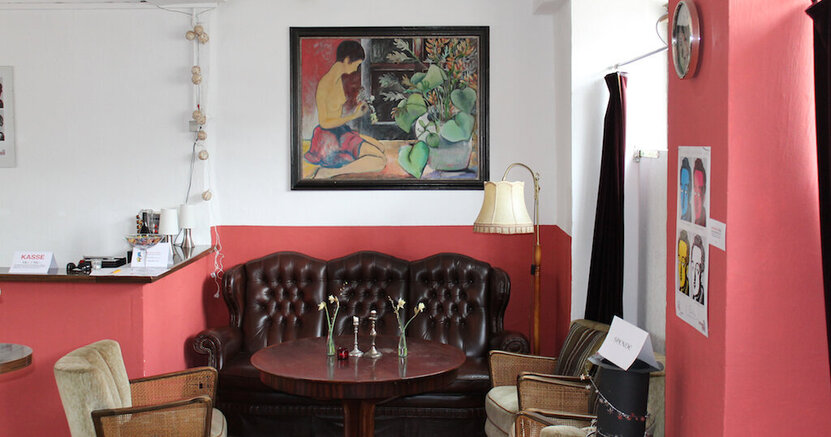 Im Theatercafé der BüchnerBühne steht an der Wand ein gemütliches Sofa, davor ein runder Tisch und drumherum einige Sessel. An der Wand hängt ein großes Gemälde.