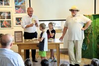 Auszählung bei der ersten Riedstädter Dschungelwahl mit Bürgermeister Marcus Kretschmann, Wahlhelfern und Forscher Dr. Cornelius Gitterbaum. Auf einem Tisch steht ein Schild mit der Aufschrift "Riedstädter Dschungelwahl". Hinter dem Tisch steht Bürgermeister Marcus Kretschmann und hält einen Wahlzettel hoch. Vor und hinter dem Tisch stehen ein Junge und ein Mädchen und schauen ihn an. Kulturbüroleiter Marco Hardy hat als Forscher Dr. Gitterbaum einen Tropenhelm auf und stützt sich seitlich auf den Tisch.