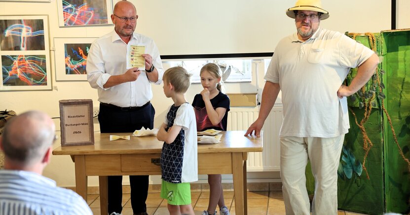 Auszählung bei der ersten Riedstädter Dschungelwahl mit Bürgermeister Marcus Kretschmann, Wahlhelfern und Forscher Dr. Cornelius Gitterbaum. Auf einem Tisch steht ein Schild mit der Aufschrift "Riedstädter Dschungelwahl". Hinter dem Tisch steht Bürgermeister Marcus Kretschmann und hält einen Wahlzettel hoch. Vor und hinter dem Tisch stehen ein Junge und ein Mädchen und schauen ihn an. Kulturbüroleiter Marco Hardy hat als Forscher Dr. Gitterbaum einen Tropenhelm auf und stützt sich seitlich auf den Tisch.