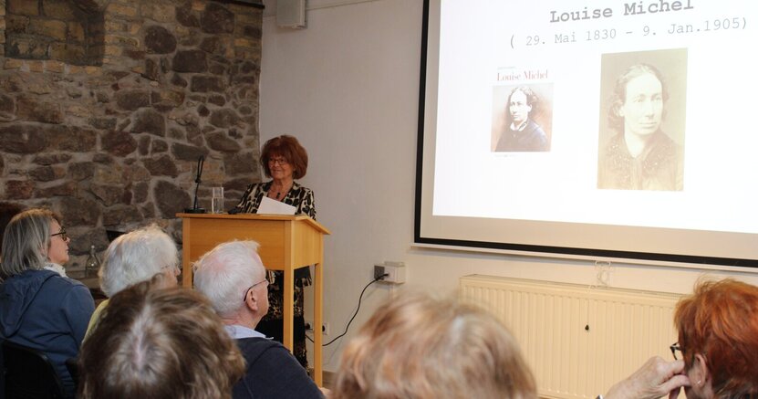 Suzanne Bohn steht in der Kunstgalerie am Büchnerhaus hinter einem Stehpult aus Holz. Auf der Leinwand rechts hinter ihr sieht man zwei Porträts von Louise Michel und dazu die Auskunft, dass sie vom 29. Mai 1830 bis zum 9. Januar 1905 gelebt hat.