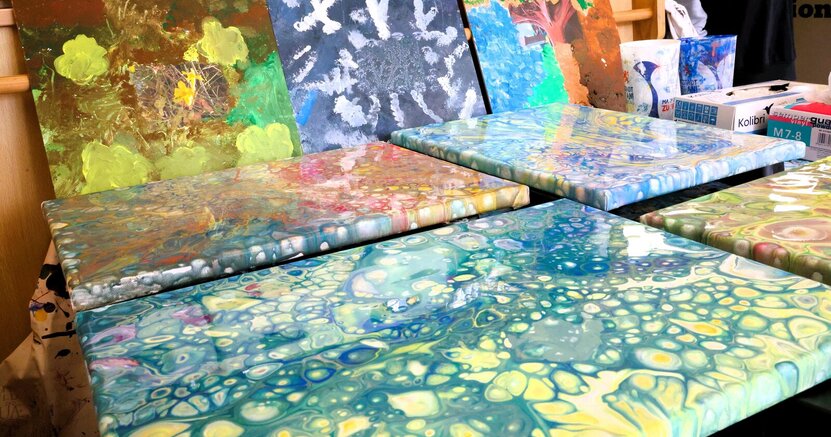 Bunte, glänzende Bilder in Mamoriertechnik liegen auf einem Tisch aus. Dahinter lehnen Bilder an einer Stütze. Bei genauem Hinsehen sieht man, dass sich in der Mitte ein Foto befindet. Das Motiv darauf wurde auf dem Bild weitergemalt.