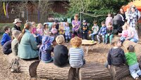 Die Kinder des Naturkindergartens sitzen mit ihren Betreuern auf querliegenden Baumstämmen, die zu einem Rund gelegt wurden. Sie klatschen und singen.