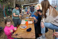 An einem langen Tisch draußen basteln Kinder und Erwachsene mit Wollfäden und Perlen. Im Hintergrund sind Bäume zu sehen.