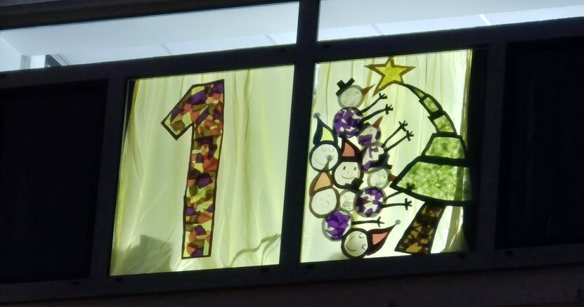 Im ersten Adventsfenster des Rathauses leuchtet eine bunte 1. Daneben ist ein Weihnachtsbaum mit Stern auf der Spitze zu sehen. Außerdem Schneemänner und Weihnachtswichtel.