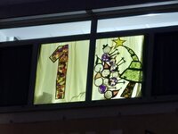 Im ersten Adventsfenster des Rathauses leuchtet eine bunte 1. Daneben ist ein Weihnachtsbaum mit Stern auf der Spitze zu sehen. Außerdem Schneemänner und Weihnachtswichtel.