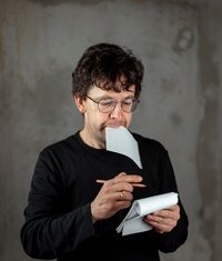 Zu sehen ist Uli Höhmann. Er hat einen Block in der einen Hand, einen Stift in der anderen Hand. Zugleich beißt er in einen Zettel..