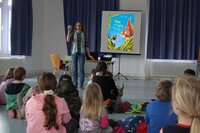 Zu sehen ist Büchereileiterin Anja Stark vor einer Leinwand, auf der ein Bilderbuch zu sehen ist. In der Hand hält sie einen Ball. Vor ihr sitzen viele Kinder auf dem Boden.