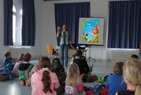 Büchereileiterin Anja Stark steht vor Kindern, die auf dem Boden sitzen. Auf der Leinwand hinter ihr ist ein Bilderbuch zu sehen.