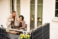 Zwei Frauen befinden sich auf einem Balkon und beugen sich über einen aufgeklappten Laptop. Außen am Balkon sind Solarpaneele angebracht.