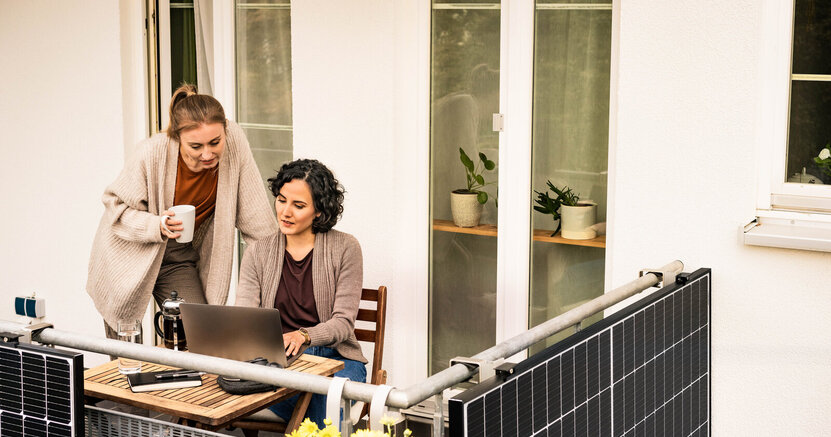 Zwei Frauen befinden sich auf einem Balkon und beugen sich über einen aufgeklappten Laptop. Außen am Balkon sind Solarpaneele angebracht.