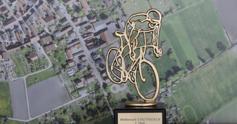 Zu sehen ist der Wanderpokal in Form eines Radfahrers.  Im Hintergrund ist ein Foto von oben von Straßen und Feldern zu sehen.